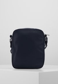 Kompaktowa torebka crossbody w ciemnogranatowym kolorze z regulowanym paskiem i zapięciem na zamek, stojąca na białym piedestale na gładkim tle.