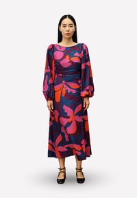 Frau in einem langärmeligen, knöchellangen Kleid mit kräftigen pinken, roten und lila Blumenmustern, die mit entspannten Händen an den Seiten steht.