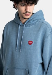 Sudadera azul clara de tela suave, con un bolsillo delantero, capucha con cordones y un pequeño parche en forma de corazón rojo con "Carhartt WIP."