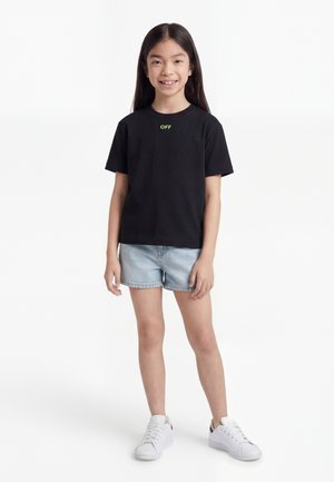 Κορίτσι που στέκεται και χαμογελά, φορώντας μαύρο μπλουζάκι με το κείμενο "OFF", ανοιχτόχρωμα τζιν σορτς και λευκά αθλητικά παπούτσια σε λευκό φόντο.