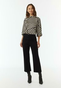 Zwart en crèmekleurige gestreepte blouse met brede mouwen, gecombineerd met zwarte high-waisted pants en enkellaarsjes, op een model tegen een effen achtergrond.