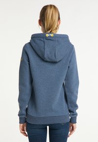 Osoba s blond vlasem v culíku, oblečená v modré mikině s kapucí a tmavých džínách, viděná zezadu na bílém pozadí.