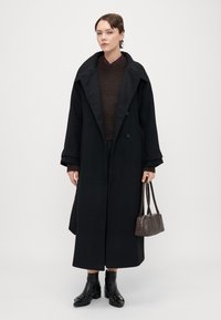 Melnas vilnas mētelis ar lielu iegriezuma apkakli, dubultās rindas dizains, apvienots ar brūnu adītu džemperi, melniem potīšu zābakiem un mazu brūnu rokas somu.