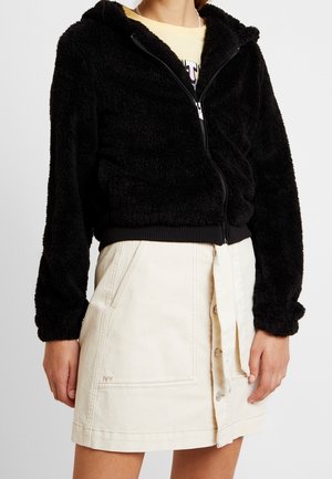Vrouw die een zwarte, pluizige jas met rits draagt over een lichtgele top en een crèmekleurige rok met knopen aan de voorkant, zakken en een taillekoord.