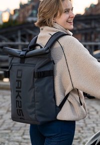 Mochila gris de tipo roll-top con correas ajustables, bolsillo lateral con cremallera y gran logotipo. El material parece duradero y resistente a las inclemencias del tiempo.