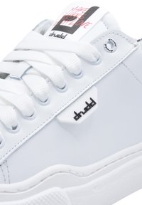 Białe sneakersy z gładką skórzaną cholewką, gumową podeszwą i okrągłymi sznurówkami. Zawierają czarny akcent logo na białej metce przy języku.