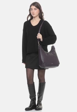 Femme portant un pull noir, une jupe, des collants et des bottes hautes tenant un grand sac à bandoulière en cuir violet foncé.
