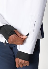 Balta vandeniui nepralaidi striukė su juodais vidiniais rankogaliais. Savybė - užtrauktuku užsegamos kišenės ir logo. Artimas plano kadras, kuriame matyti ranka, reguliuojanti rankovę, kad būtų patogu ir tinkamai priglustų.