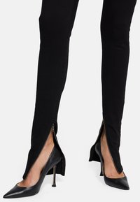 Czarne wąskie spodnie z bocznymi zamkami, wykonane z gładkiego, elastycznego materiału, o dopasowanym kroju, zestawione z czarnymi, spiczastymi szpilkami.