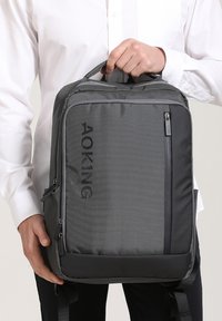 Sac à dos en tissu gris avec une forme structurée, doté d'une poche zippée à l'avant, d'une impression de logo et d'une poignée supérieure. Bretelles réglables.
