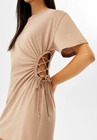 Krótka beżowa sukienka typu t-shirt z marszczonymi bokami i sznurowanym detalem, z okrągłym dekoltem i krótkimi rękawami, wykonana z miękkiej, lekkiej tkaniny.