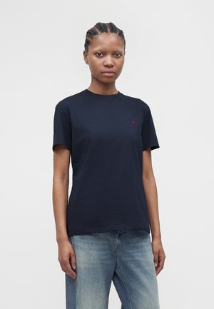 Tânără cu părul împletit, purtând un tricou simplu negru cu mâneci scurte și blugi albastru deschis, stând în fața unui fundal simplu.