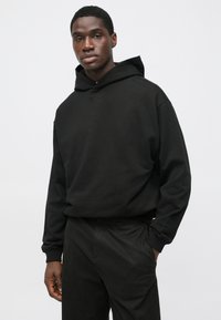 Čierna mikina s kapucňou z mäkkej tkaniny s voľným strihom, vybavená sťahovacou kapucňou a nenápadným logom "BOSS" na hrudi. Kombinovaná s čiernymi nohavicami.