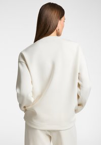 Felpa oversized color crema con una vestibilità rilassata, spalle scese e una texture liscia. Presenta un orlo e polsini a costine.