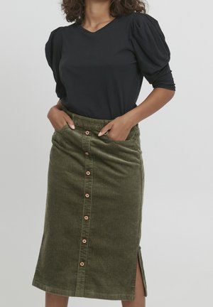 Femme portant un haut noir à manches bouffantes et une jupe midi en velours côtelé vert olive boutonnée avec une fente latérale, mains dans les poches.