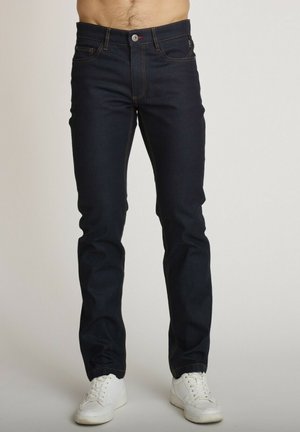 Homme portant un jean slim bleu foncé et des baskets blanches debout devant un fond uni.