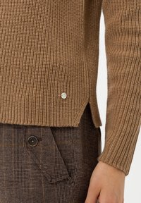 Pull en maille marron côtelée avec fentes latérales, ornée d'un petit accent argenté. Associé à un pantalon à carreaux marron avec un détail de bouton sur la poche.