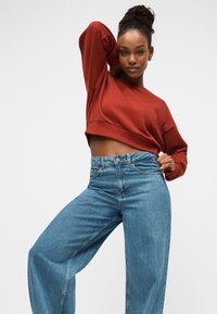 Κοντή φουτεράδα σκουριασμένου χρώματος με μακριά μανίκια, συνδυασμένη με ψηλόμεσο, ανοιχτό μπλε παντελόνι με φαρδιά μπατζάκια. Λείο ύφασμα, casual εφαρμογή, ελάχιστα μεταλλικά στοιχεία.