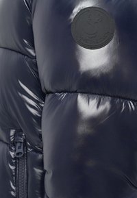 Shiny navy blue puffer jacket with zipper and round black patch featuring a duck silhouette and "SAVE THE DUCK" text.