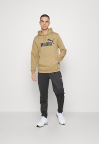 Felpa beige con logo Puma nero, abbinata a pantaloni jogger neri. L'outfit presenta una tasca frontale e scarpe da ginnastica bianche casual.
