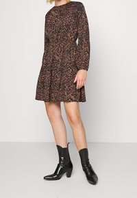 Robe noire avec motif floral en rose et or, manches longues, taille froncée et jupe évasée, associée à des bottines noires en cuir à motif crocodile.