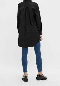 Zwarte button-up shirt met een rugplooi, lange mouwen, gebogen zoom, gedragen over blauwe skinny jeans en zwarte schoenen. Effen textuur.
