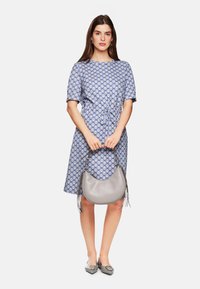 Blau gemustertes Kleid mit kurzen Ärmeln, tailliert, kombiniert mit einer geschwungenen grauen Handtasche mit Fransenakzenten und spitzen Ballerinas.