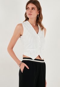 Bela brezrokavna majica z ozkimi vertikalnimi črnimi črtami, V-izrezom, sprednjimi gumbi in dvema stranskima žepoma. Nosijo jo z črnimi hlačami.