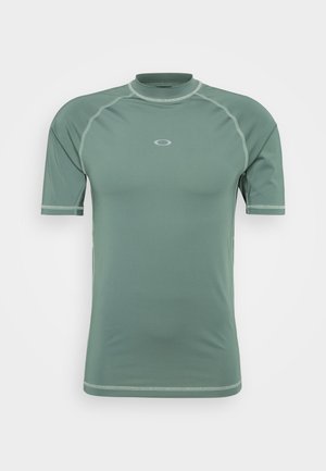 Kortermet skjorte i dempet grønn stoff med en jevn tekstur, med høy krage og kontrasterende trim. Diskret logo sentrert på brystet.