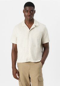Camisa de botones de manga corta en color beige con tela texturizada, dos bolsillos frontales y diseño de cuello tipo campamento. Combinada con pantalones cargo beige.