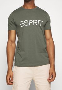 Olivgrünes Baumwoll-T-Shirt mit kurzen Ärmeln und Rundhalsausschnitt, mit dem weißen "ESPRIT"-Logo, das über die Brust gedruckt ist. Glatte Textur.