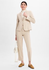 Beige op maat gemaakte blazer met twee knopen, bijpassende slim fit broek en beige loafers. Witte handtas met een lange schouderband. Glide stoftextuur.