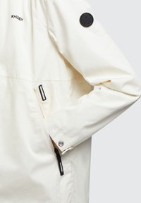 Jachetă bej deschis cu o textură netedă. Are un buzunar cu fermoar, un ecuson circular negru pe mânecă și mansete reglabile.