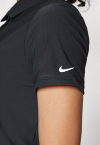 Čierna polokošela Nike s rebrovaným golierom a bielym logom na ľavom rukáve. Tkanina vyzerá hladko a prispôsobene.