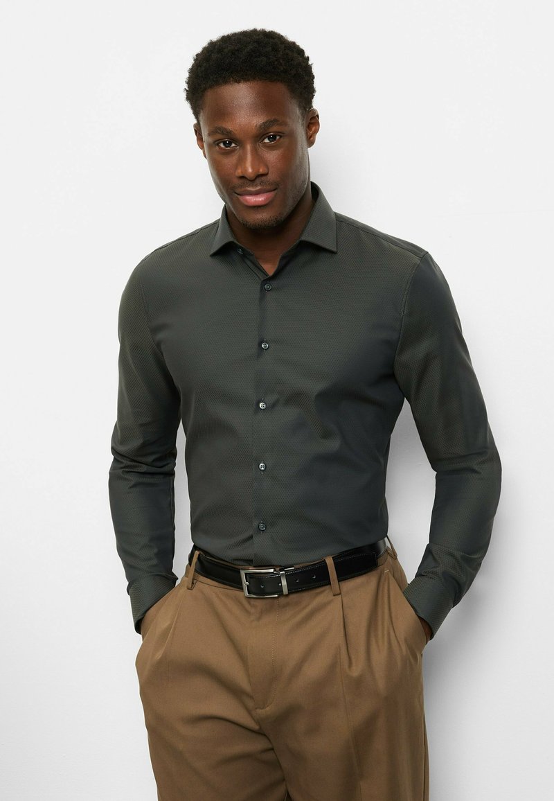 Dunkelgrünes, gemustertes Hemd mit Kragen und durchgehender Knopfleiste, kombiniert mit braunen Hosen und schwarzem Gürtel; glatte Stofftextur.