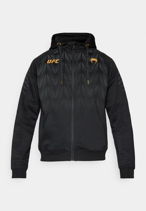 Hoodie noir à fermeture éclair avec un design géométrique, comprenant des accents orange, des cordons de serrage et des poignets et ourlet côtelés. Fabriqué dans un tissu doux et texturé.