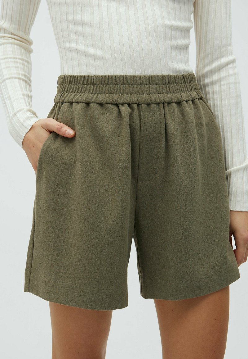 Personne portant un short taille élastique vert olive avec poches latérales et un haut blanc à manches longues côtelé, main dans la poche gauche.