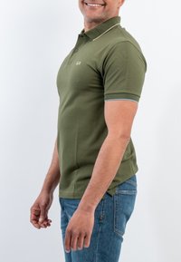 Polo verde oliva con maniche corte, bordo blu a contrasto ai polsini delle maniche e logo sottile sul petto. Indossato con jeans blu.