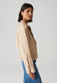 Giacca beige cropped realizzata in tessuto liscio, con colletto, due tasche frontali e polsini elastici con dettaglio a bottone.