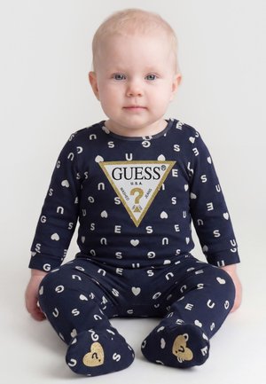 Lyshåret baby, der sidder og har en mørkeblå Guess-dragt med hvide bogstaver og hjerte-mønstre, guldt logo på brystet og fødderne.