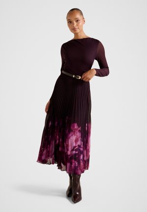 Γυναίκα με μακρυμάνικο σκούρο μωβ φόρεμα με πλισέ φούστα που έχει ροζ λουλουδάτο μοτίβο, μαύρη ζώνη και σκούρα καφέ μπότες, στέκεται μπροστά σε λευκό φόντο.