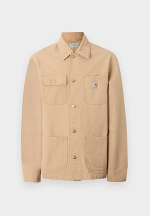 Veste Carhartt beige avec quatre poches avant, fermeture par boutons, et écusson du logo de la marque sur la poche poitrine droite.