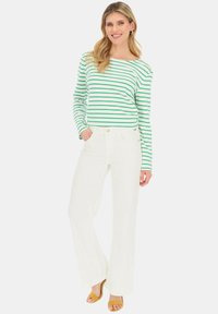 Długi rękawowy top w zielono-białe paski, zestawiony z białymi spodniami o szerokich nogawkach. Modelka nosi żółte sandały, prezentując swobodny strój.