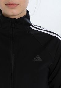 Sort zip-up jakke med høj krave, der har tre hvide striber på skuldrene og et diskret sort Adidas logo på brystet. Glat tekstur.
