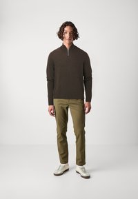 Suéter marrón con cremallera en el cuello, pantalones verdes y zapatillas blancas. La tela es suave; los pantalones tienen un corte recto y una textura sutil. Diseño minimalista.