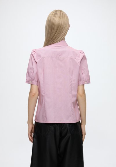 Femme aux longs cheveux blonds portant un t-shirt rose à manches courtes et un pantalon noir, vue de dos sur un fond uni.