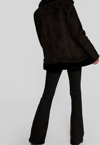 Chaqueta de ante sintético negra con cuello, mangas largas y un ajuste ligeramente holgado, combinada con pantalones negros de campana. Textura suave.