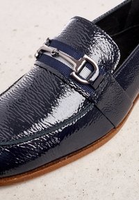 Navy patent leather loafer with a textured finish, featuring a silver buckle accent and a woven striped band. Flat brown sole.