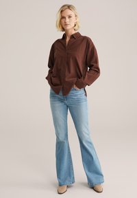 Blonde woman wearing a loose brown button-up shirt, light blue flared jeans, and beige pointed shoes, standing with hands in pockets.