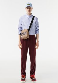 Bolso cruzado beige con logo, gorra azul clara, camisa de botones a rayas, pantalones burdeos y zapatillas multicolor con cordones rojos.